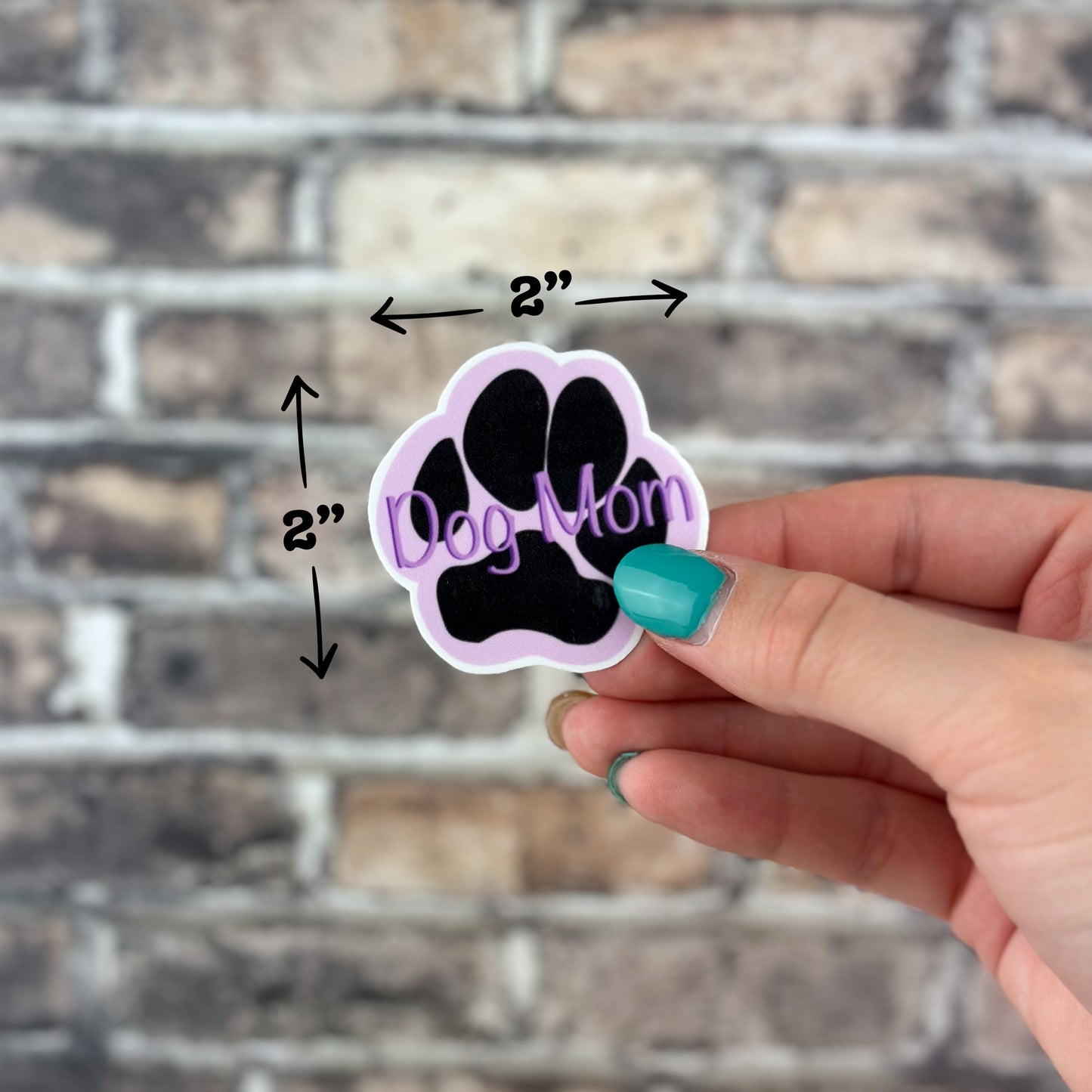 A hand featuring teal nail polish grips a decorative sticker designed as a black paw print with a white outline. The phrase "Dog Mom" is elegantly inscribed in purple on the paw, enhancing its allure. Positioned against a brick backdrop, the Dog Mom Purple Sticker measures 2 inches by 2 inches.