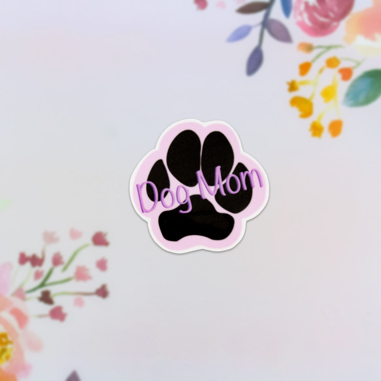 The Dog Mom Purple Sticker features a black paw print and the words "Dog Mom" in pink, set against a pastel backdrop adorned with floral designs in shades of pink, yellow, and green. This sticker adds charm to any surface.