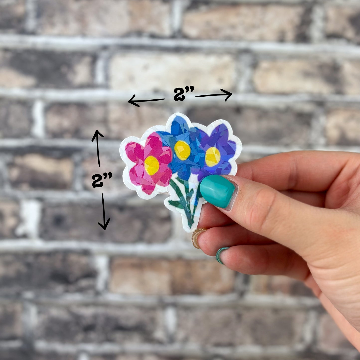 A hand with turquoise nails displays the Flower Bouquet Holographic Sticker against a textured brick wall background, with dimensions of 2 inches by 2 inches.