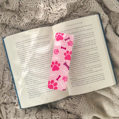 A Pink Paws Bookmark, featuring delightful paw prints and the words "woof" and "paw," is gently placed on the open pages of a book. This ideal reading accessory lies on a knitted blanket, creating a cozy ambiance for any book enthusiast.