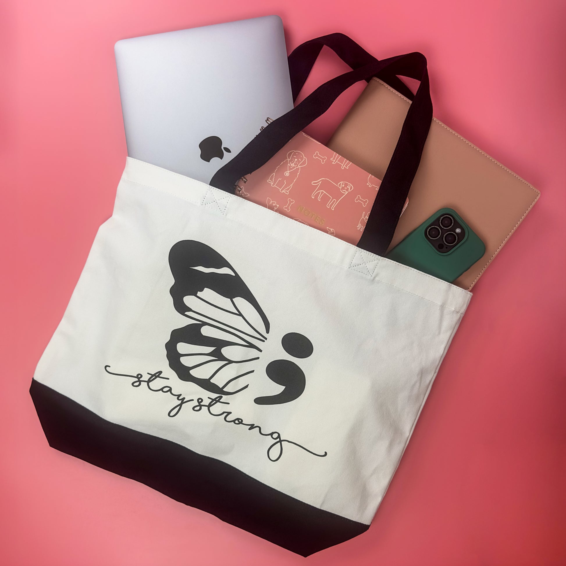 The Stay Strong Butterfly Tote Bag, made from eco-friendly cotton canvas, is beautifully displayed on a pink backdrop. Featuring a butterfly design alongside the words "stay strong," it stylishly holds a laptop, a pink animal-themed notebook, and a smartphone while raising awareness for suicide prevention.