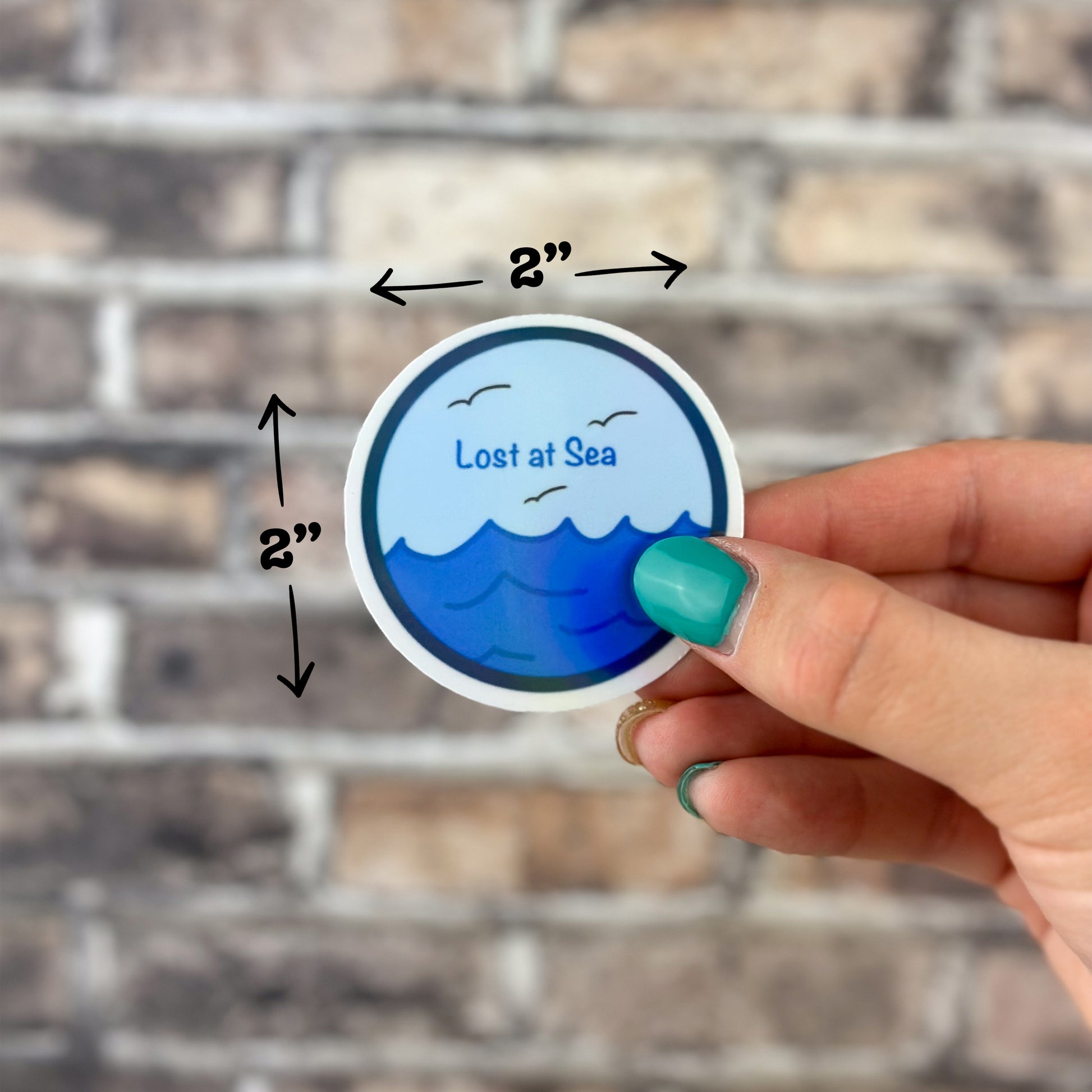 A hand with teal-painted nails is holding a Lost at Sea Holographic Sticker against a brick wall. The sticker features a blue sea design with the text "Lost at Sea," measures 2 inches in diameter, and is dishwasher-safe. Arrows highlight its dimensions.