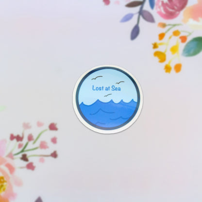 This dishwasher-safe sticker, named "Lost at Sea," features a holographic design with an ocean theme, including waves and birds. The text "Lost at Sea" is prominently displayed in the center against a vibrant background of colorful floral patterns, creating a captivating fusion of nature's elements.