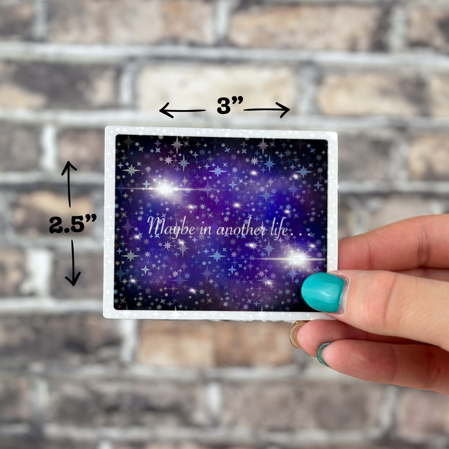 A hand with turquoise nail polish holds a small card against a brick wall background. The card, measuring 3 inches by 2.5 inches, features a starry sky design along with the "Maybe in Another Life… Holographic Sticker.