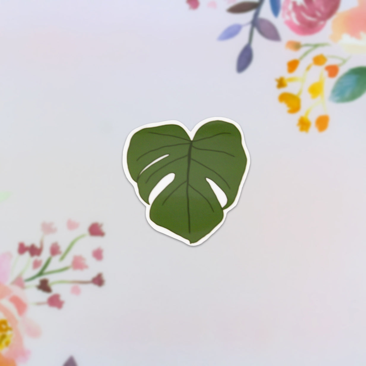 Centered on a soft, pastel floral background, the Monstera Leaf Sticker is adorned with surrounding flowers in pink, orange, and purple hues—ideal for plant enthusiasts wanting to add a natural touch.