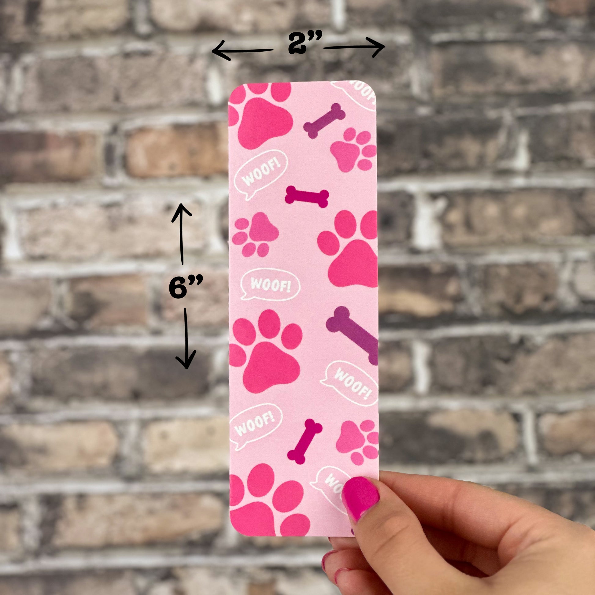 A hand holds the Pink Paws Bookmark—a delightful essential for every book enthusiast. Adorned with endearing dog-themed motifs including paw prints, bones, and speech bubbles displaying "WOOF!", this bookmark measures 2x6 inches and serves as the ideal reading accessory showcased against a brick wall background.