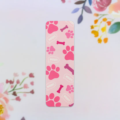 The Pink Paws Bookmark, an essential accessory for book enthusiasts, features a delightful pink background adorned with vivid paw prints, purple bones, and lively white "woof!" speech bubbles. Set against a subtle floral backdrop, it makes for an ideal whimsical reading companion.