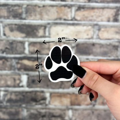 With black nail polish, an individual holds a Puppy Paw Sticker measuring approximately 2 inches by 2 inches, set against a brick wall backdrop—a perfect decorative piece for dog enthusiasts.