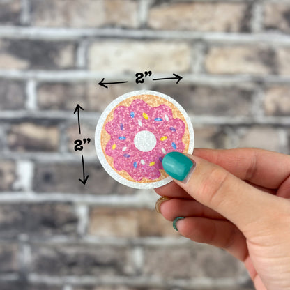 A hand with turquoise nail polish presents the Strawberry Donut Holographic Sticker, highlighting its decorative allure. This 2-inch round sticker features a pink-frosted donut adorned with sprinkles and shines vibrantly against a brick wall setting. Arrows indicate its dimensions, emphasizing that it is both fashionable and dishwasher safe.